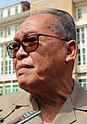 A 3/4 profile image of Kong Korn. He has short grey hair and tan skin, he is wearing a gray shirt and glasses with a brown tint.