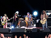 A band performs on a stage while several audience members raise their arms up. In the background, two spotlights flash brightly.