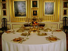 A round table covered with white table cloth and set for six with silverware and a centrepiece, standing in front of a wall covered with yellow and white striped wallpaper and framed paintings