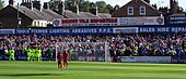 One of the stands of the Bootham Crescent association football ground, with supporters cheering and players standing on a grass field below