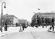 Photographie en noir et blanc d'une place traversée par des piétons et voitures, au fond des bâtiments.