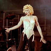 Madonna wearing a beige corset and black pants. She has blonde curly hair and has a headset microphone to her mouth