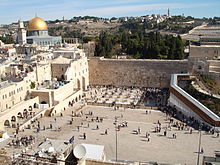 A large open area with people bounded by old stone walls. To the left is a mosque with large golden dome.