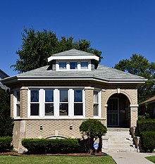 A light beige brick building with a three-sided bay to the viewer's left and a small corner porch to the viewer's right and a single dormer in the center, with a lawn that contains a bush and a small tree immediately in front of the bay.