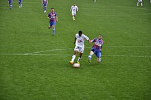 Joueur en train de tirer vers le but, un défenseur essayant de le contrer en le taclant par le côté.