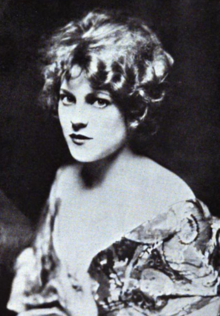 A young white woman with curly light hair, wearing a low-cut print dress against a dark background