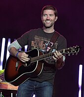 A dark-haired young man in a t-shirt and jeans playing a guitar