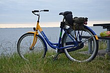 Farbfoto. Blau-gelbes Hollandrad, tiefer Einstieg, Beleuchtung, Gepäckträger, keine Handbremse, keine Gangschaltung. Auf dem Rahmen steht „OV-fiets.nl“. Im Hintergrund ein Gewässer.