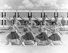 Four female and four male cheerleaders wearing striped sweaters and making a choreographed pose in front of the new ASC library
