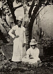 Une femme se tient debout, l'autre est assise. Elles portent une robe blanche et un chapeau typiquement victorien.