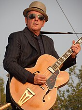 A color photograph of Elvis Costello, who is playing a guitar outside.