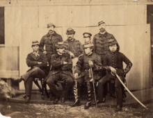 Photographie sépia de huit officiers prenant la pose, quatre au premier rang assis sur un banc et les quatre autres debout derrière eux.