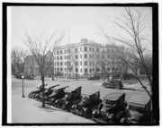 A black-and-white photograph of a street scene. The focus is a small, light-colored five story hotel with the first floor below grade, rounded turrets at the corners and an ornamental parapet, viewed catty-corner looking southwest across the intersection of New Jersey Avenue and C Street. The trees are bare and the street is lined with over a dozen twenties-era automobiles.