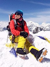 Die Farbfotografie im Hochformat zeigt Wang Jing im Schnee sitzend. Sie trägt eine gelbe Schneehose und eine dicke rote Winterjacke. Ihre Mütze ist blau und sie trägt eine Sonnenbrille. Im Hintergrund sind schneebedeckte Berge erkennbar.