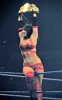 A dark-haired, tanned Caucasian female straddles the top rope of a wrestling ring with blue ropes. She is wearing a red patterned crop top, and matching shorts and kneepads. She is holding a wrestling championship above her head with both hands.