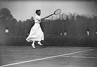 Женщина во всей белой одежде бьет справа теннисной ракеткой в ​​правой руке, это черно-белая фотография.