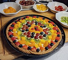 A fruit pizza on a kitchen counter and cutting board, the fruit pizza surrounded by bowls of fruit pieces that were used as toppings for the pizza.