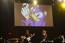 A Japanese man (left) plays a guitar while an American man (right) sings in front of a crowd.