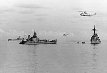 The stern of a destroyer, floating in calm water. The bow has been removed completely. Two intact warships are close by, one to the right of frame, the other behind and partially obscured by the floating stern, and two helicopters are flying overhead.