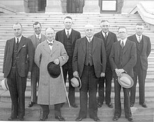 Eight middle aged white men, seven wearing three piece suits and one wearing an overcoat that obscures what he's wearing beneath, stand in two rows in front of some steps.