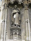 Stone statue of a standing man wearing robes and a mitre carrying a tablet in one hand and holding his other hand up.