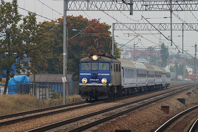 Na zdjęciu widać zbliżający się do widza pociąg z wagonami przedziałowymi na przedzie którego jedzie niebieska lokomotywa z włączonymi lampami. Obok kolejne tory, nad nią przewody elektryczne.