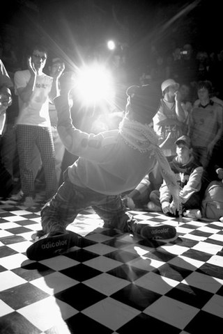 A close-up black and white photo of a male hip-hop dancer surrounded by a small crowd in a nightclub while performing on a checkerboard dance floor.