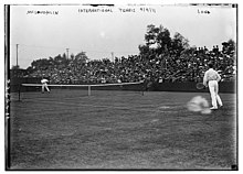 A black and white picture of two players in white pants and shirts playing on a grass court in front of a small crowd