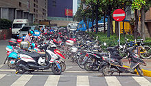 Two rows of motorbikes, many showing their age and use, parked next to a city street corner. There is a large white-bar-on-red-circle "do not enter" sign at the upper right.