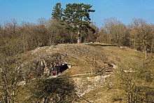 Photographie en couleurs d'une colline percée de deux ouvertures, recouverte d'un tapis herbeux, d'arbres et de roches affleurantes, un étroit sentier aménagé d'un banc visible à mi-hauteur.