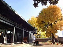 Foto a colori. Entrata di tempio dai muri laccati neri ombreggiato da un albero dal fogliame giallo con il cielo blu.