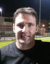 A man wearing a black T-shirt with a soccer training field in the background.