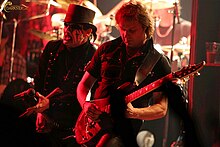 Two members from the band King Diamond are shown at a concert performance. From left to right are the singer and an electric guitarist. The singer has white and black face makeup and a top hat. Both are wearing black.