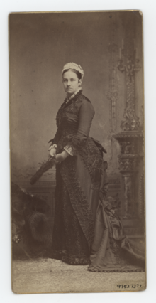 A full body, black and white photograph of a stern looking woman in a dark coloured dress.