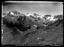 Schwarz-Weiß Fotografie einer Berglandschaft mit Felsen und Schnee. Etwa in der Bildmitte sieht man relativ klein ein Gebäude aus Stein mit reflektierendem Dach. Es steht an einem Abhang, ein Schotterpfad führt auf das Gebäude zu.