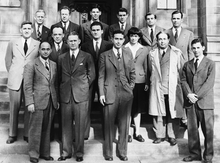 A group of people in suits standing in three rows on the steps in front of a stone building.