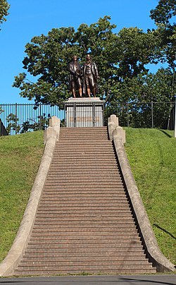 Photograph showing a long, broad flight of steps leading to a large metal statue of two men. There are about 50 steps. The two men in the statue are roughly life-sized, and are standing on a large stone pedestal. The pedestal has bronze plaque that reads 