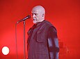 Vor einem roten Hintergrund steht der Sänger Peter Gabriel aufrecht an seinem Mikrofon; Hemd und geschlossener Blazer sind schwarz, die Hände hinter dem Rücken verschränkt, das Gesicht wirkt nachdenklich