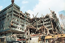 Seen from ground level, a heavily damaged and partially collapsed seven story building is surrounded by debris