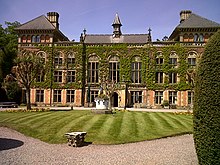 The frontage of a house with a three-storey central block, and two, four-storey, towers at either end. The house is faced in red brick, and is covered with ivy. A neatly-mown lawn stands in front.