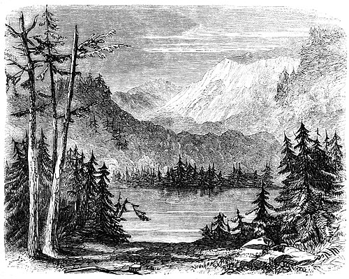 alt = A small lake, surrounded on every side by rocky shores, evergreen trees, and steep barren mountains. In the foreground, evergreen trees on both sides frame a glimpse of the lake; in the background are high peaks with snow on their tops.