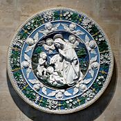 A circular terracotta plaque, sculptured in relief and glazed in intense colours of blue and green with white figures and motifs. At the centre the Virgin Mary, watched by John the Baptist, kneels in adoration of the baby Jesus. Little cherubs look on