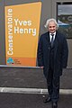 Photo d'un homme posant devant un bâtiment ; panneau portant son nom « Yves Henry ».