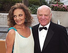 A woman with wavy and curly hair wearing a white dress lined with a teal fabric smiles as she looks to her right while standing next to a man wearing a tuxedo