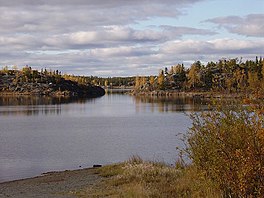 A small body of water at the base of a beach in the foreground, with a passage between two high, stony areas and more land behind it. All the land is covered in forest with a mix of deciduous and evergreen trees.