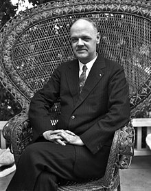 Photograph of a Caucasian man, dressed in a three-piece suit, and sitting in a wicker chair