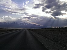 Photographie prise depuis la route, qui n'a pas de marquage. On voit un paysage de la steppe de Kouraï, avec des montagnes aux cimes enneigées au fond. Mais le est ennuagé, les nuages ne laissant que peu passer la lumière, créant une atmosphère sombre.