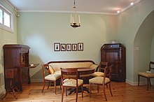 Farbfotografie eines Innenraums mit einer Holzcouch und Holzstühlen aus beigem Polster um einen runden Tisch. An der hinteren, hellgrünen Wand hängen fünf Porträts im Schattenriss. An beiden Ecken stehen ein Sekretär und ein Schrank. Von der Decke hängt ein einfacher Kronleuchter. Links ist an der oberen Wandseite ein kleines Fenster und rechts ist eine Wandnische.