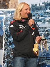 A blonde woman, wearing a predominately black jacket and blue jeans, stands in front of a large poster of a winter landscape, smiles and looks to the right. She holds a microphone in her right hand and several gold medals in her left hand.
