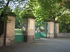 Zdjęcie przedstawia bramę wejściową parku. Brama składa się z 4 wysokich słupów, pomiędzy którymi znajdują się zielone bramki. Przed bramą znajduje się wybrukowany kostką plac.